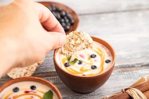 Yoghurt with bilberry and caramel in clay bowl on gray wooden, hand dipping cookies in yogurt, side