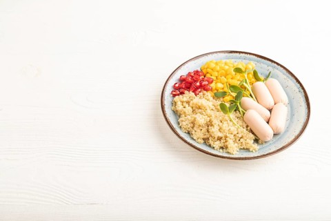 Mixed quinoa porridge, sweet corn, pomegranate seeds and small sausages on white wooden background.