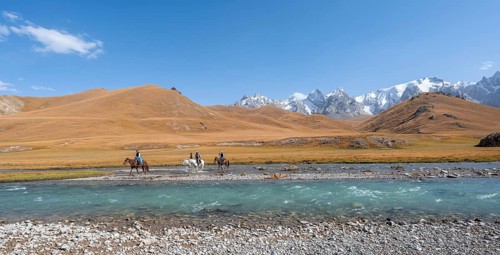 Riders on horses on the river bank, mountain landscape with yellow meadows and river Kol Suu,