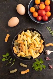 Rigatoni colored raw pasta with tomato, eggs, spices, herbs on black concrete background. Top view,