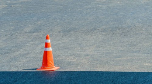 Orange traffic cone sitting in a pave parking lot on a sunny day with a shadow of a building in