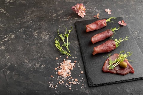 Slices of smoked salted meat with green pea microgreen on black concrete background. Side view,