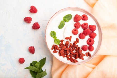 Yogurt with raspberry and goji berries in ceramic bowl on white concrete background and orange