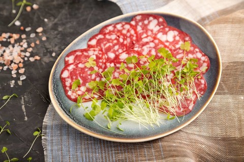 Slices of smoked cervelat salami sausage with spinach microgreen, salt and pepper on black concrete
