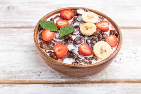 Chocolate cornflakes with milk and strawberry in wooden bowl on white wooden background. Side view,