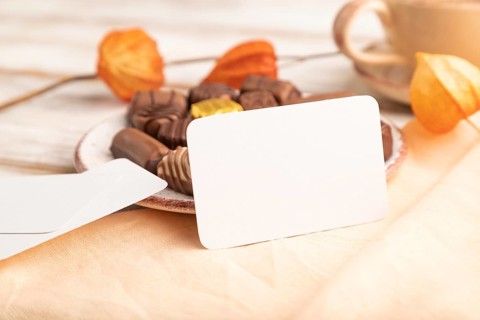 White business card with chocolate candies and physalis flowers on a white wooden background and