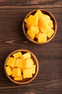 Dried and candied mango cubes in wooden bowls on brown wooden textured background. Top view, flat