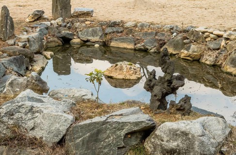 Piece of basalt shaped like tree next to small man made pond surrounded by granite boulders in