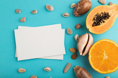 White paper business card with ripe cut papaya, orange, seashells on blue pastel pastel background.