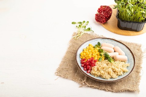 Mixed quinoa porridge, sweet corn, pomegranate seeds and small sausages on white wooden background.