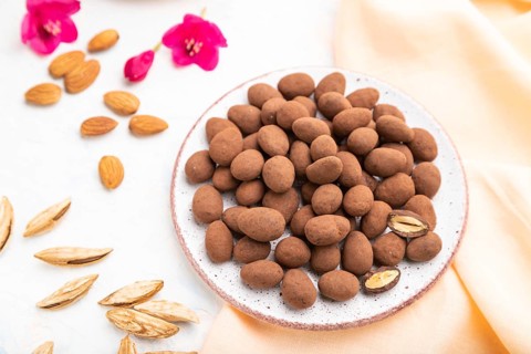 Almond in chocolate dragees on ceramic plate and a cup of coffee on white concrete background and