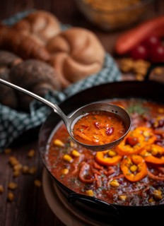 Soup ladle scooping bean soup with sweetcorn and peppers from a pot, basket with pastries in the