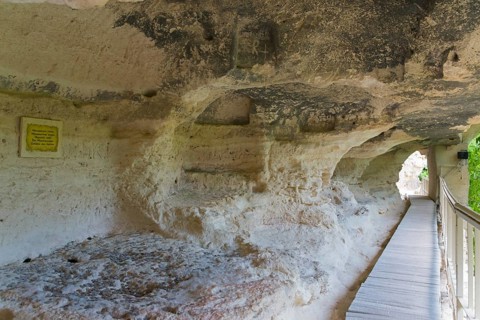 A walkway leads through a cave entrance, accompanied by information signs, cells of the monks,