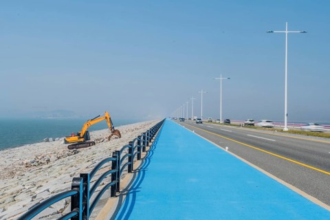 Backhoe sitting on ocean causeway next to blue concrete sidewalk on divided highway with speeding