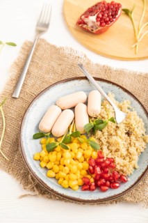 Mixed quinoa porridge, sweet corn, pomegranate seeds and small sausages on white wooden background.