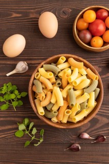 Rigatoni colored raw pasta with tomato, eggs, spices, herbs on brown wooden background. Top view,
