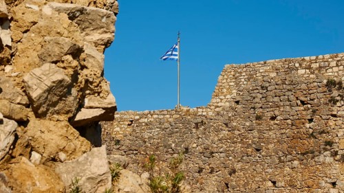 Part of an old fortress wall with a waving Greek flag and blue sky, Chlemoutsi, High Medieval