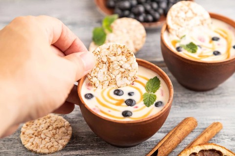 Yoghurt with bilberry and caramel in clay bowl on gray wooden, hand dipping cookies in yogurt, side
