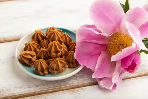 Homemade soft caramel fudge candies on blue plate on white wooden background, peony flower