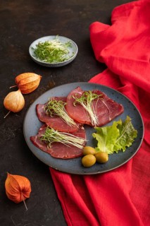 Slices of smoked salted meat with cilantro microgreen on black concrete background and red textile.