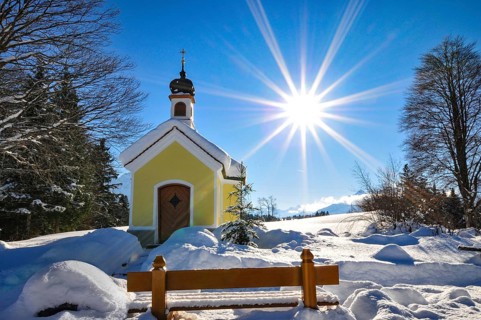 Backlit photograph of the snow-covered Maria Chapel on the humpback meadows of Werdenfelser Land