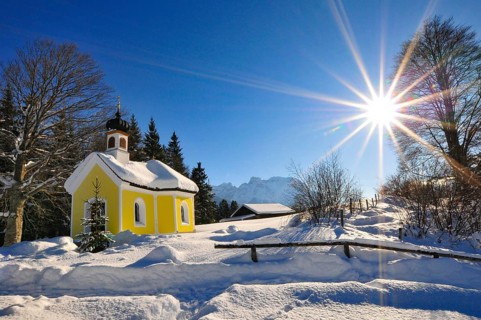 Backlit photograph of the snow-covered Maria Chapel on the humpback meadows of Werdenfelser Land