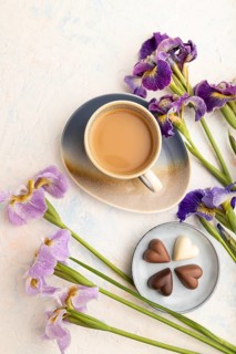 Cup of cioffee with chocolate candies and lilac iris flowers on white concrete background. top