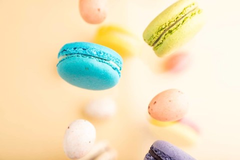 Multicolored flying macaroons and chocolate eggs frozen in the air on blurred beige background. top