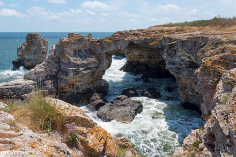 Rugged rock formations and natural arch on a stormy coast against a bright blue sky, rock arch,