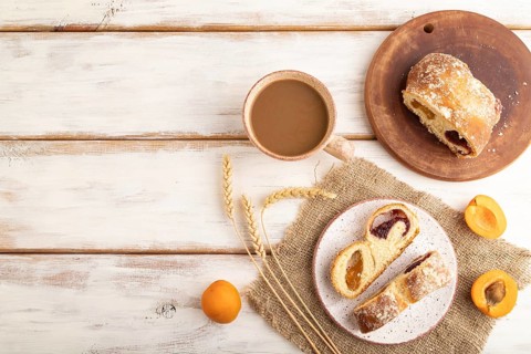 Homemade sweet bun with apricot jam and cup of coffee on white wooden background and linen textile.