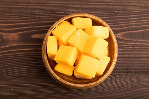 Dried and candied mango cubes in wooden bowls on brown wooden textured background. Top view, flat