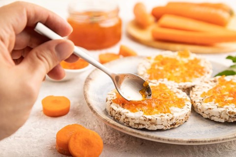 Carrot jam with puffed rice cakes on gray concrete background with hand. Side view, close up,