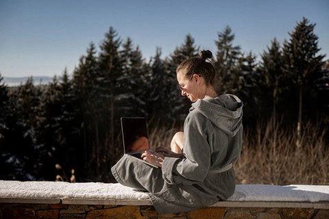 Woman in a bathrobe sits on a snowy stone wall in nature and works on her laptop