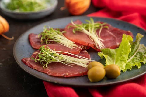 Slices of smoked salted meat with cilantro microgreen on black concrete background and red textile.