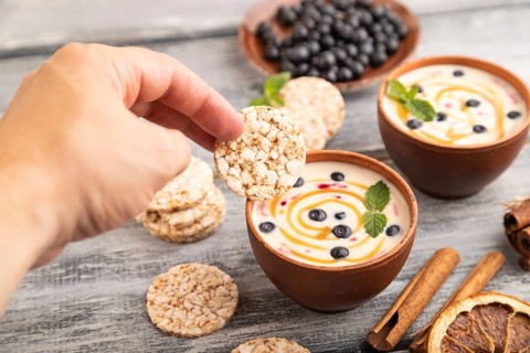 Yoghurt with bilberry and caramel in clay bowl on gray wooden, hand dipping cookies in yogurt, side