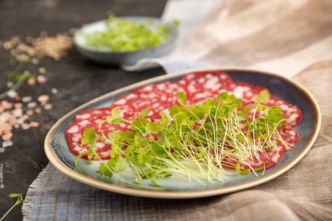 Slices of smoked cervelat salami sausage with spinach microgreen, salt and pepper on black concrete