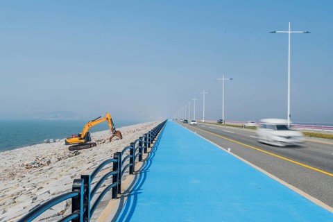 Backhoe sitting on ocean causeway next to blue concrete sidewalk on divided highway with speeding