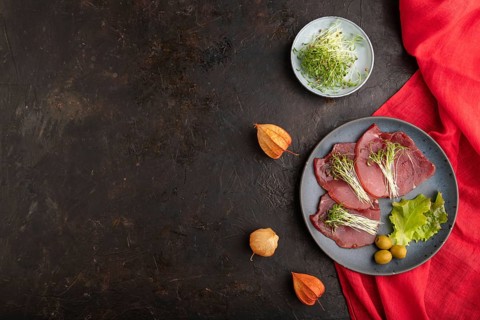 Slices of smoked salted meat with cilantro microgreen on black concrete background and red textile.