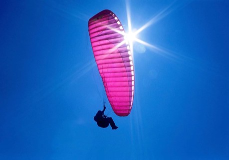 A purple, pink paraglider flies in the blue sky, backlit by the sun and lots of sunbeams