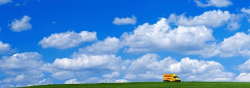 A DHL delivery van on a country road in the middle of meadows with blue sky and many white fair