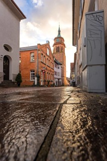 Damp cobblestones lead to the church tower between houses, graffiti on the wall in late light, Weil