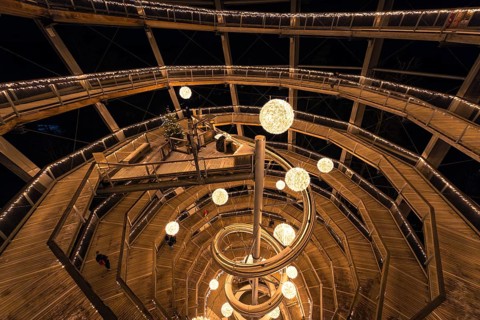 People walk through a spiral-shaped wooden construction with lights at night, magical forest