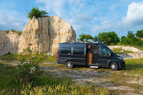 A motorhome stands in front of a rocky formation under a blue sky with clouds, mountain with