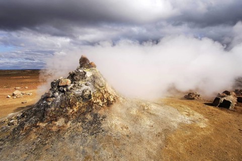 Close-up of steaming vent or fumarole in a geothermal landscape with a view of distant hills at