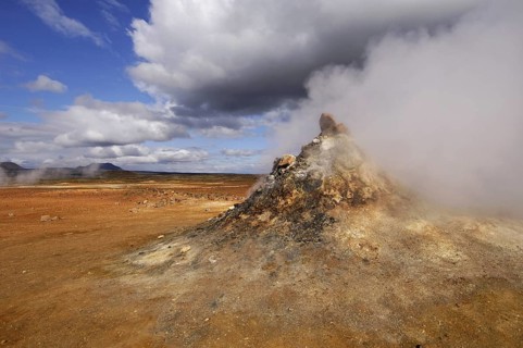 Close-up of steaming vent or fumarole in a geothermal landscape with a view of distant hills at