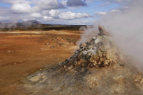 Close-up of steaming vent or fumarole in a geothermal landscape with a view of distant hills at