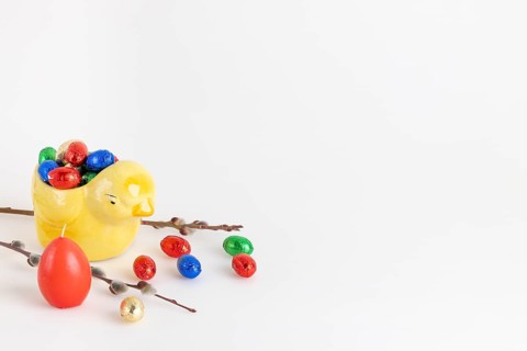 Colourful chocolate eggs in a yellow ceramic chick next to a palm kitten and candle, white