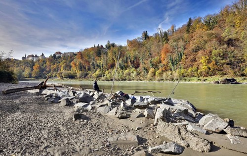 An angler sits on large stones by a river in autumn, with a castle perched on a wooded hill in the