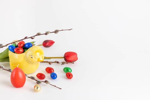 Colourful chocolate eggs in a yellow ceramic chick next to palm catkins, tulips and candle, white