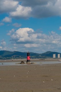 Red and black lighthouse on tidal flats during low tide with mountains and cloudy blue sky in